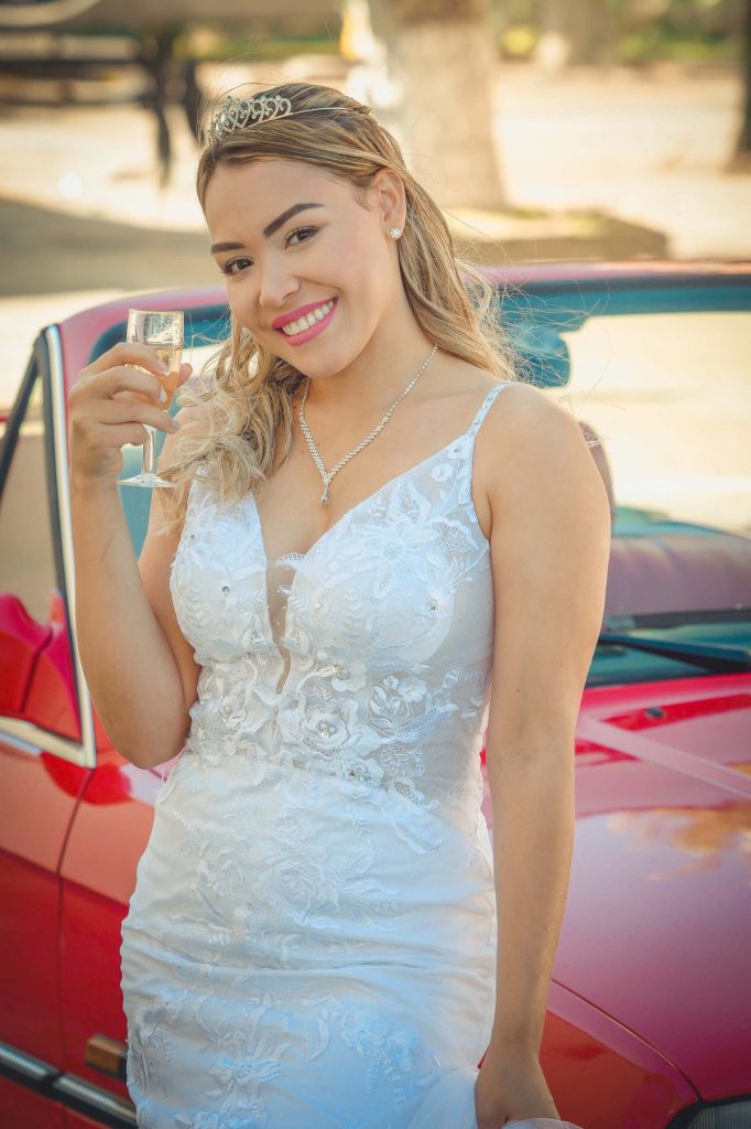 La mariée est devant une voiture rouge et elle à un verre à la main